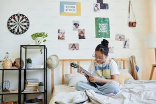 Ritratto a figura intera della ragazza adolescente asiatica che suona l'ukulele mentre è seduta sul letto in una stanza accogliente, spazio di copia