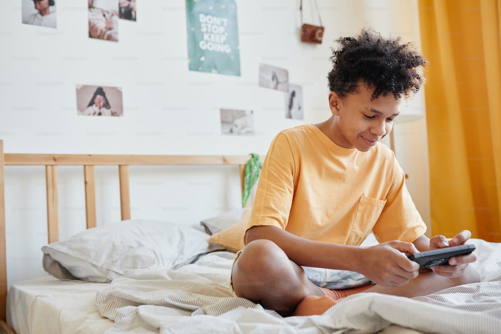 Retrato de cuerpo entero de un adolescente de raza mixta sentado en la cama y jugando juegos móviles a través de un teléfono inteligente, espacio de copia
