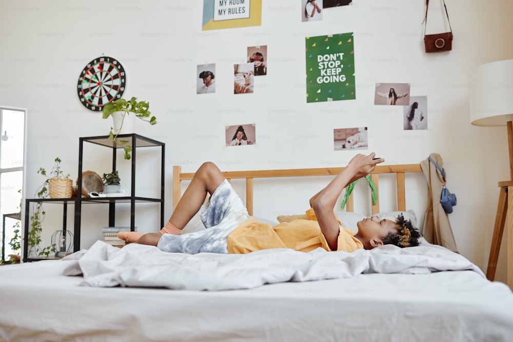 Retrato de cuerpo entero de un adolescente de raza mixta acostado en la cama y usando un teléfono inteligente, espacio de copia