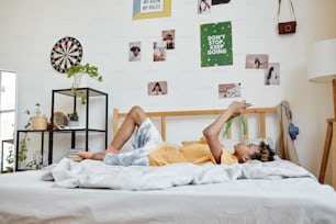 Full length portrait of mixed-race teenage boy laying on bed and using smartphone, copy space