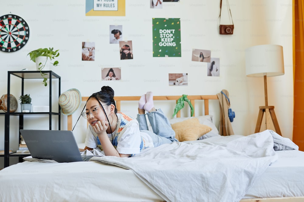 Portrait of teenage Asian girl using laptop on bed in cozy room interior, copy space