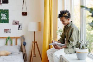 Side view portrait of mixed-race teenage boy sitting on window at home and writing in notebook, copy space