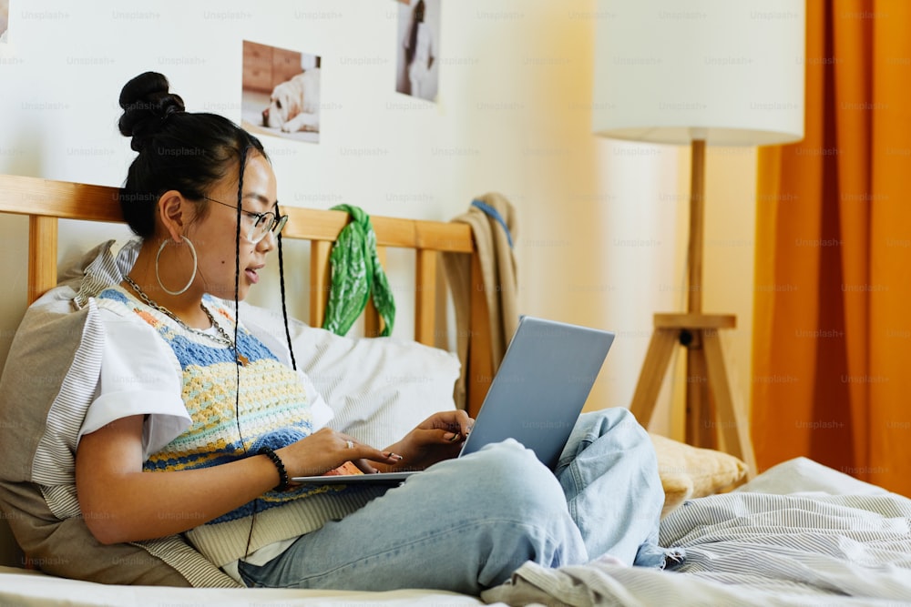 Side view portrait of teenage Asian girl using laptop while sitting on bed in cozy room interior, copy space