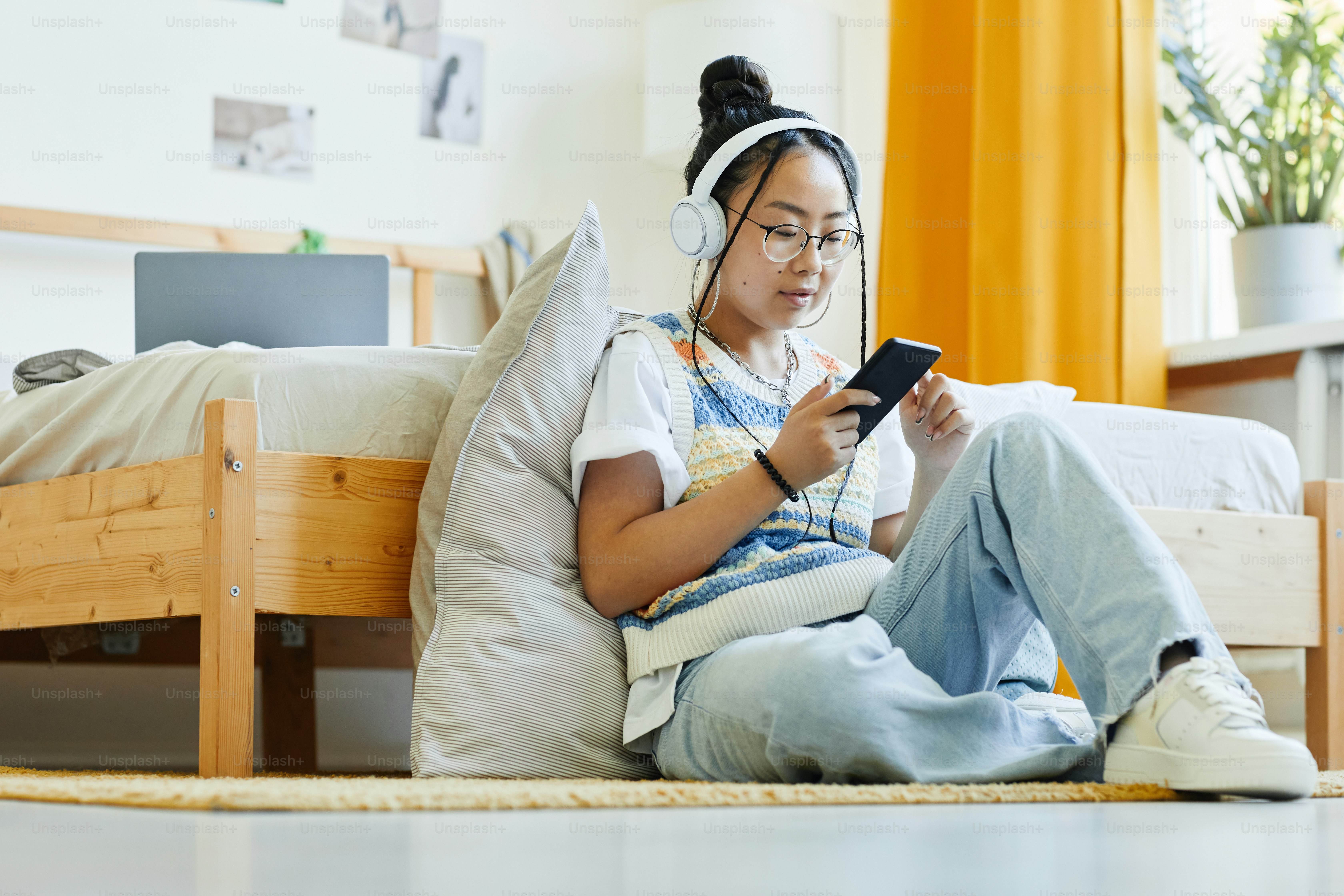 Potret panjang penuh gadis remaja trendi menggunakan smartphone dan mendengarkan musik sambil duduk di lantai di ruangan yang nyaman, ruang salinan