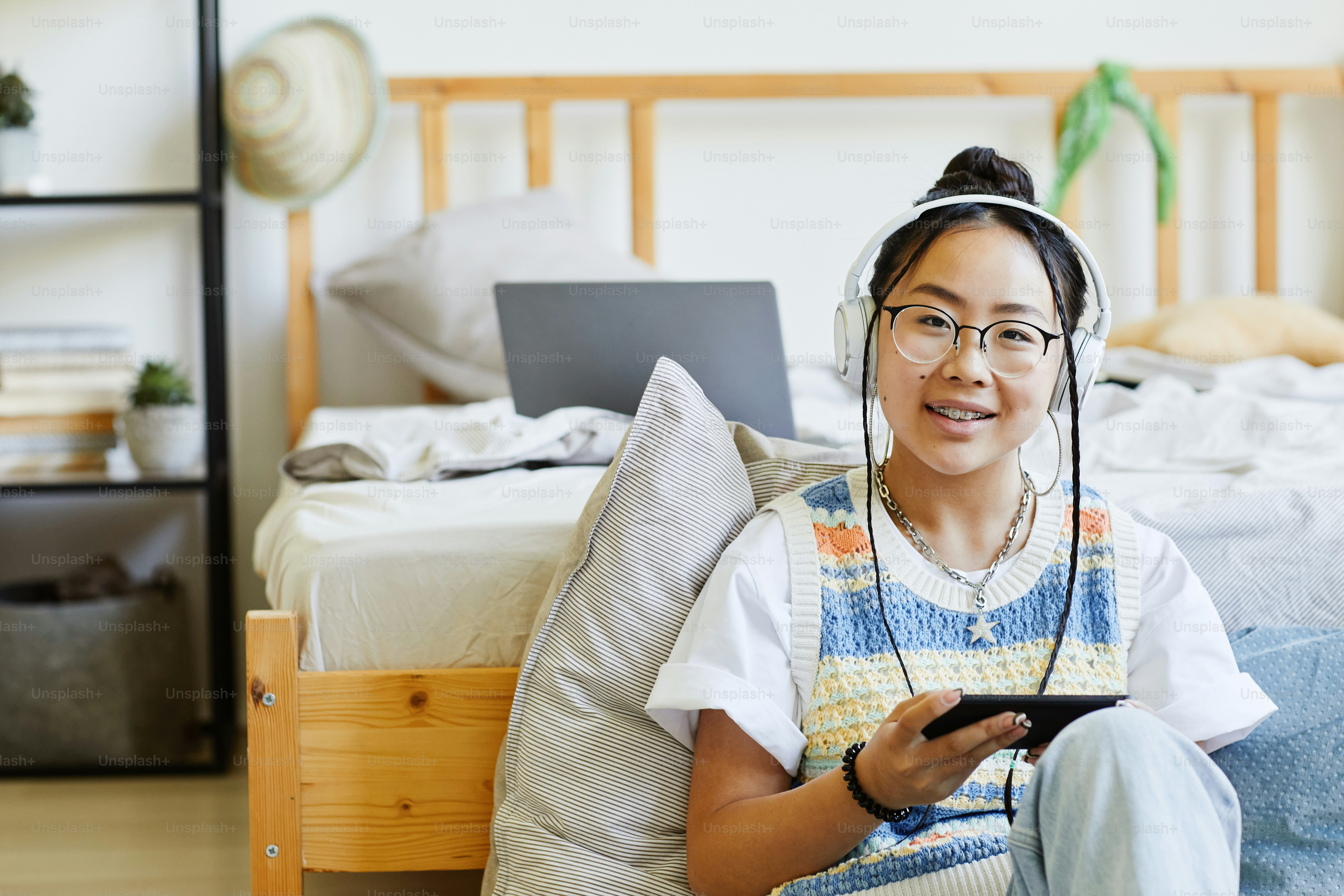 Portrait of trendy teenage girl listening to music while sitting on floor in cozy room and looking at camera, copy space