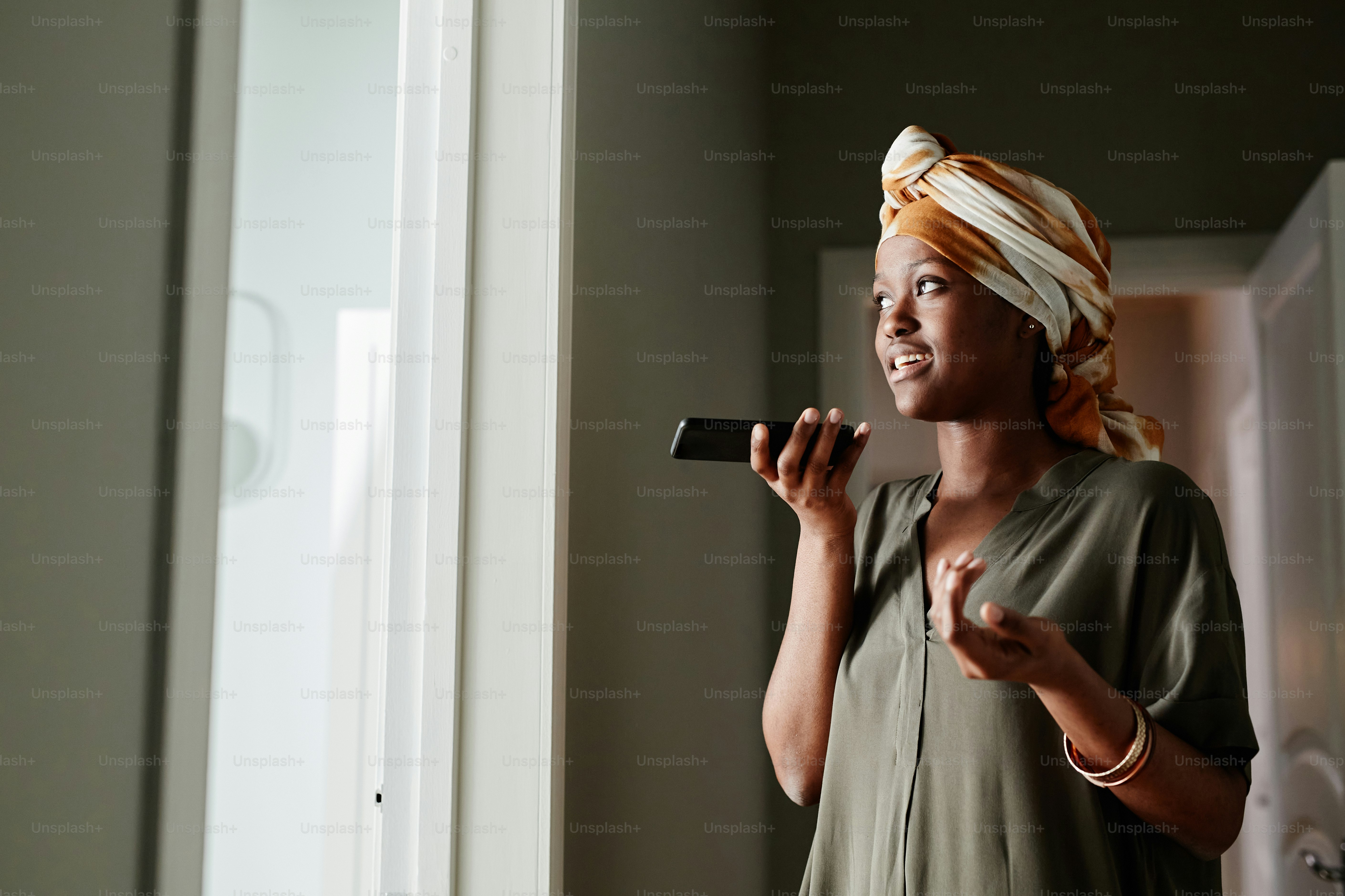 Side view portrait of young African-American woman recording voice message while standing by window at home, copy space