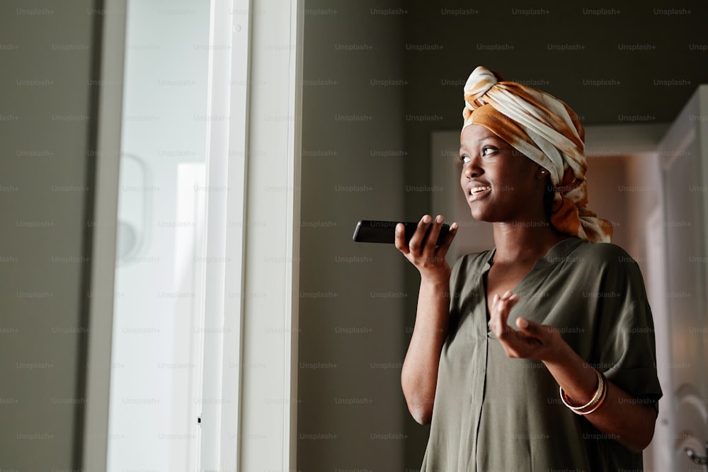 Side view portrait of young African-American woman recording voice message while standing by window at home, copy space