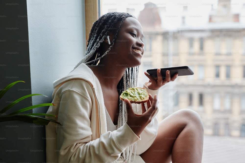 Side view portrait of young African-American woman recording voice messages while sitting by window and enjoying morning at home, copy space