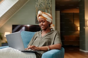 Retrato de una joven afroamericana sonriente relajándose en casa y usando una computadora portátil, espacio de copia