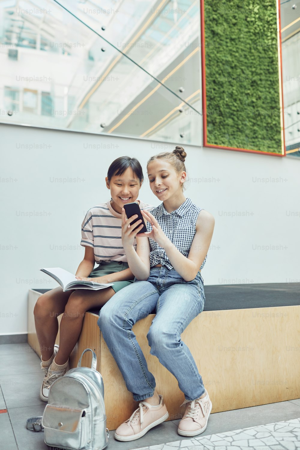 Vertical full length portrait of two schoolgirls using smartphone in school hall and smiling