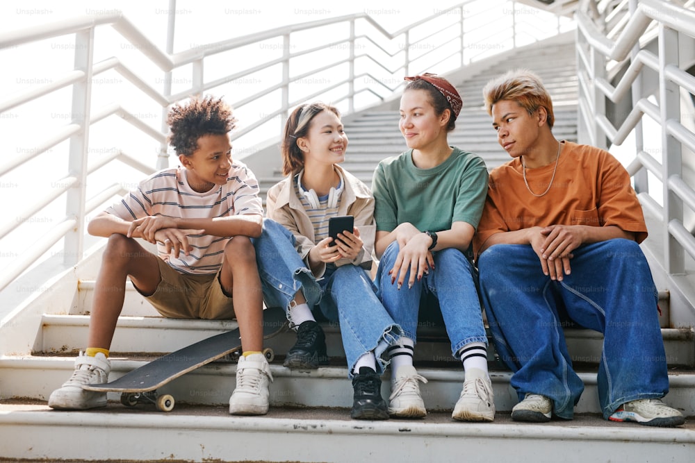 Full length portrait of diverse group of teenagers sitting on metal stairs outdoors in urban setting all smiling and chatting
