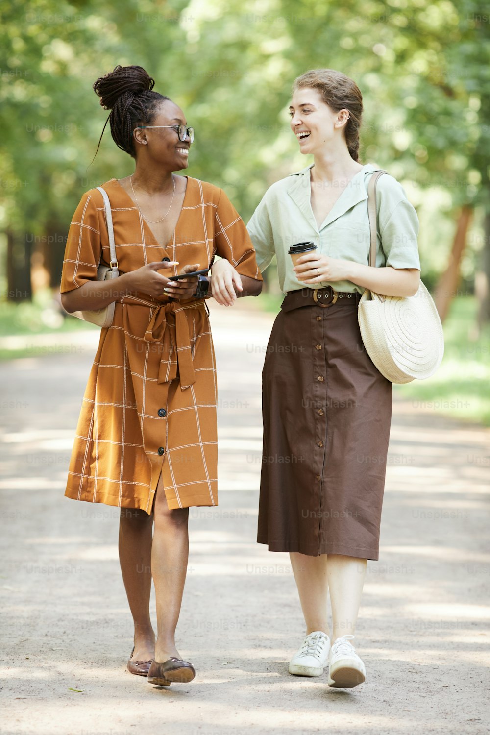 Vertical full length portrait of two smiling young women chatting outdoors while enjoying walk in park together