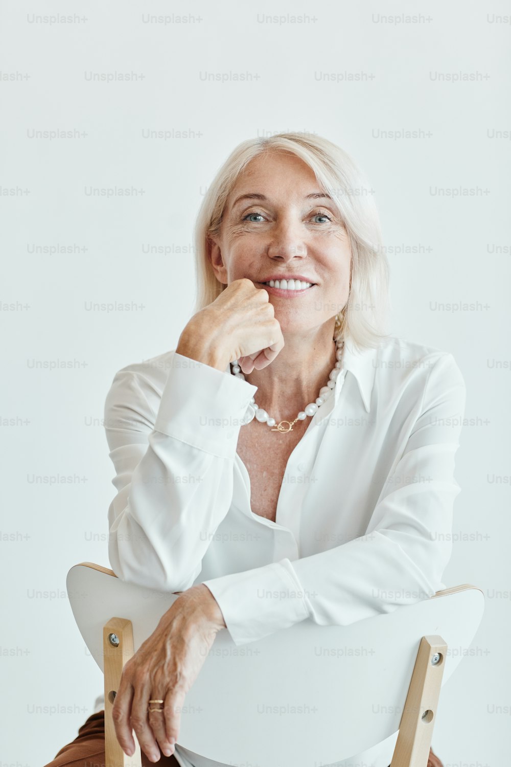 Vertical portrait of elegant mature woman smiling while sitting on chair backwards against white background