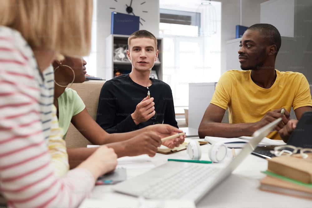 Un grupo diverso de estudiantes que trabajan juntos en la mesa de la biblioteca se centran en el joven que habla con amigos mientras estudia