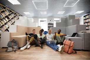 Wide angle view at diverse group of young students studying together while sitting on floor in college library, copy space