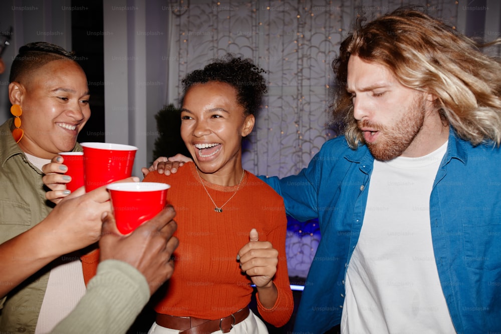 Grupo diversificado de jovens amigos desfrutando de festa em casa dentro de casa e segurando copos vermelhos, filmados com flash