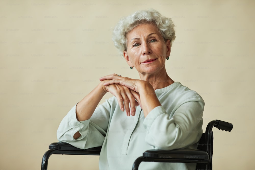 Retrato de una elegante mujer mayor mirando a la cámara en silla de ruedas sobre fondo neutro, copia espacio