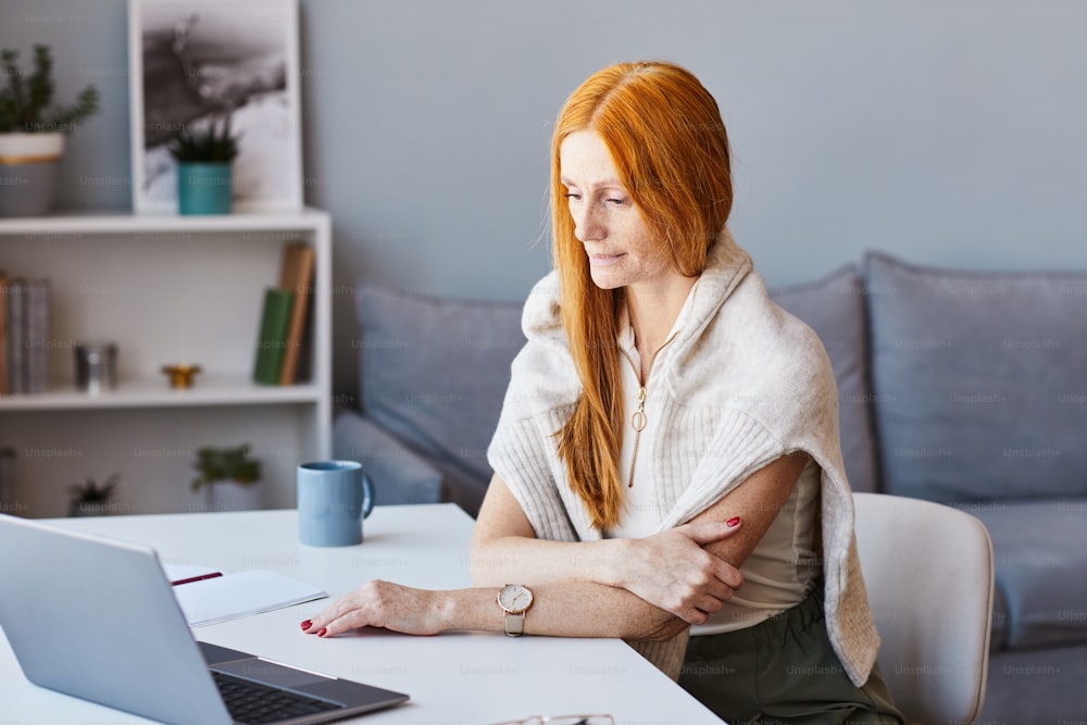 Retrato da mulher de cabelos vermelhos elegante usando o laptop enquanto trabalha no escritório em casa, espaço de cópia