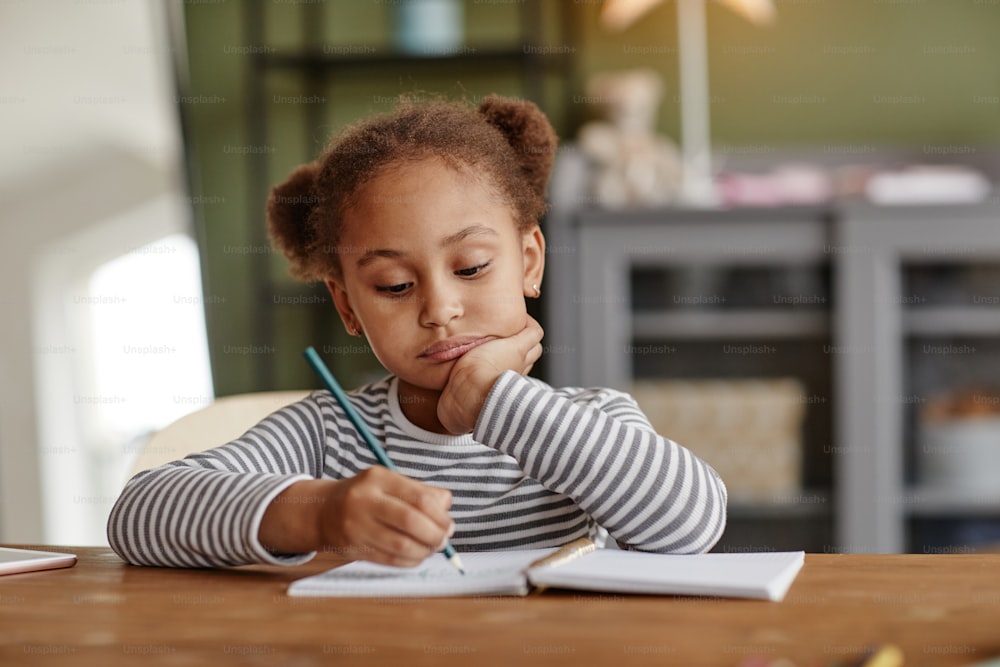 Front view portrait of cute African-American girl drawing while sitting at wooden desk, copy space
