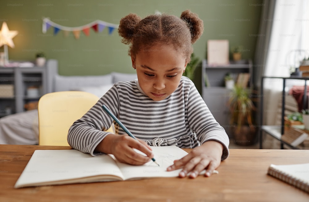 Front view portrait of cute African-American girl doing homework while sitting at wooden desk, copy space