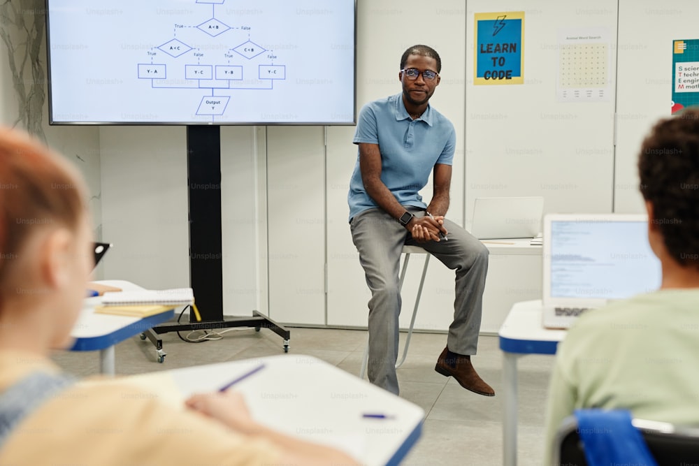 Portrait of young African-American teacher sitting on desk while teaching coding class for children, copy space