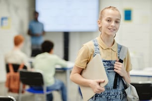 Retrato de cintura para arriba de una adolescente sonriente con mochila y mirando a la cámara en el aula de la escuela, espacio de copia