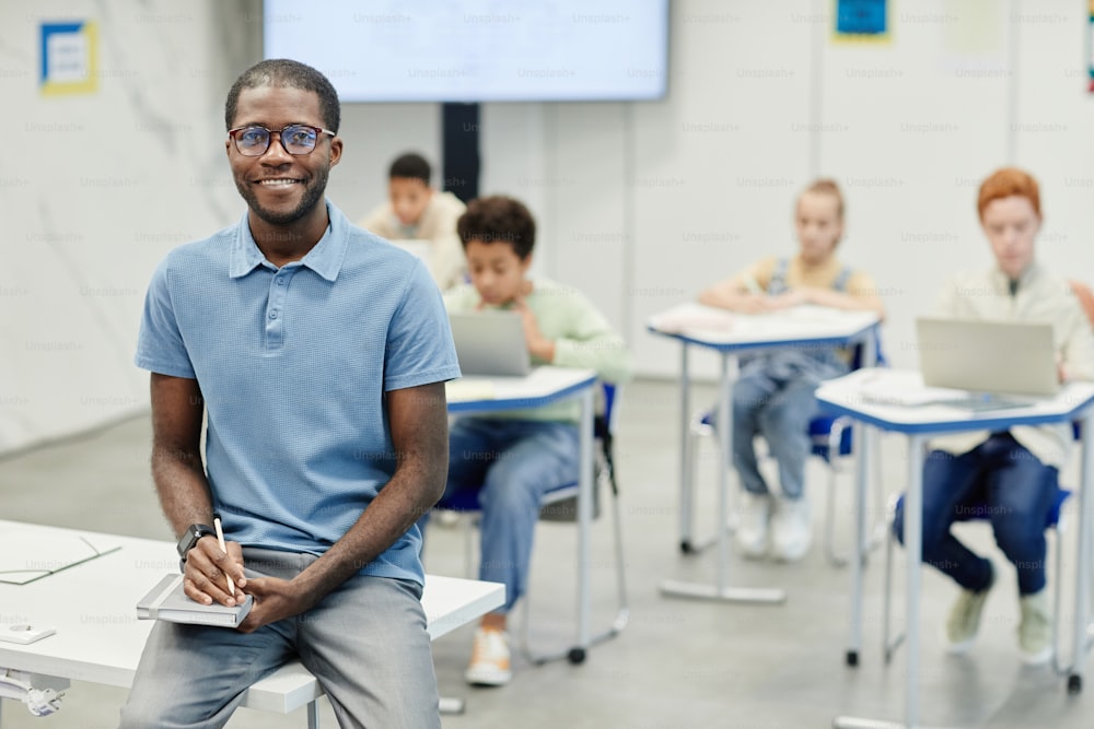 Portrait of young African-American teacher sitting on desk and smiling at camera with children in background, copy space