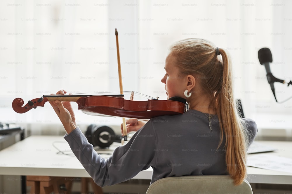 Vue arrière d’une jeune femme blonde jouant du violon dans un studio contre la lumière de la fenêtre, espace de copie