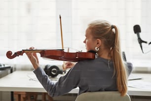 Vista trasera de una joven rubia tocando el violín en el estudio contra la luz de la ventana, copia del espacio