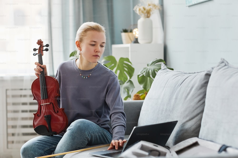 Ritratto di giovane donna bionda che suona il violino a casa e guarda la lezione online, spazio di copia