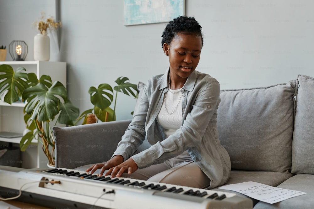 Portrait of young black woman playing synthesizer at home and composing music in cozy home setting, copy space