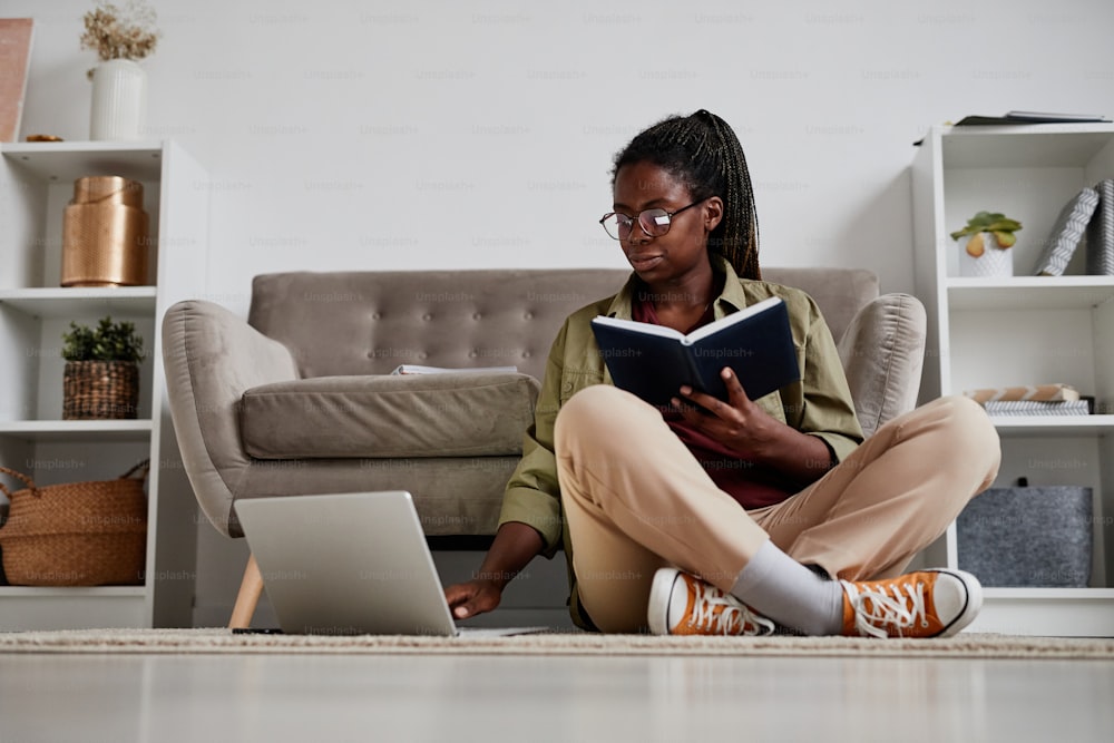 Portrait en pied d’une jeune femme afro-américaine travaillant à domicile assise par terre dans un cadre confortable, espace de copie