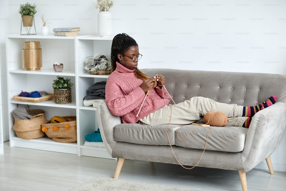 Ritratto a figura intera della giovane donna afroamericana che lavora a maglia a casa mentre si gode un fine settimana accogliente, spazio di copia