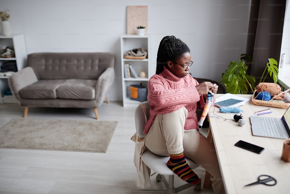 Ritratto ad angolo alto di giovane donna afroamericana che lavora a maglia a casa e guardando lo schermo del laptop, guardando video educativi, spazio di copia