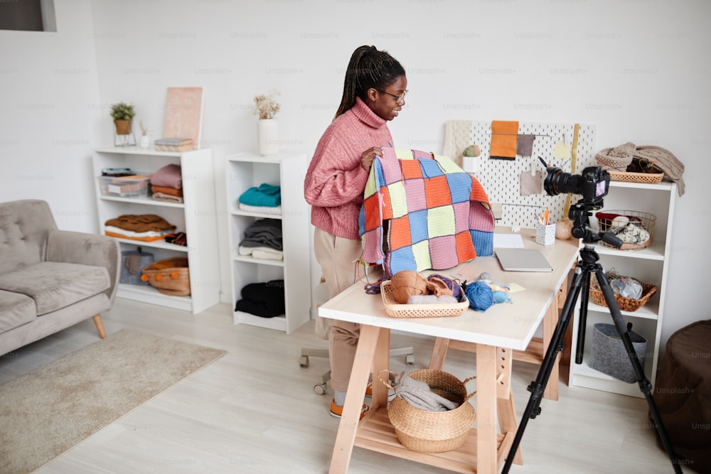 Ritratto grandangolare di giovane donna afroamericana che lavora a maglia a casa e registra video o livestream, spazio di copia