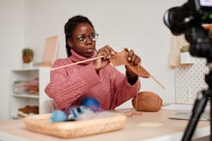 Portrait of young African-American woman knitting at home and recording video or online education class