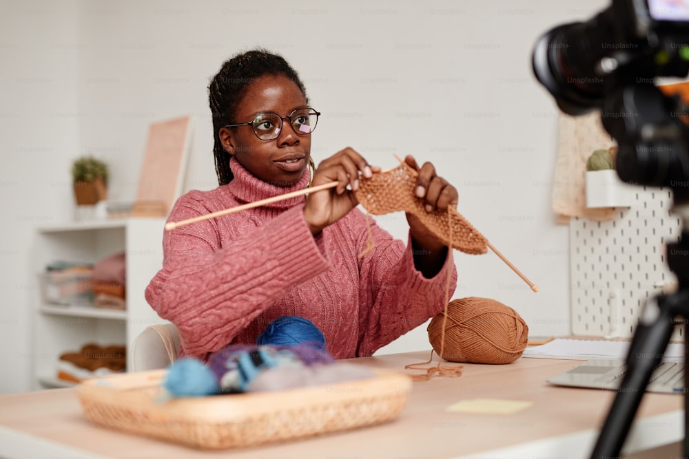 Ritratto di giovane donna afroamericana che lavora a maglia a casa e registra video o lezione di educazione online