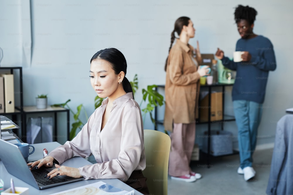 Portrait of young Asian woman using laptop at workplace with people chatting in background in company office, copy space