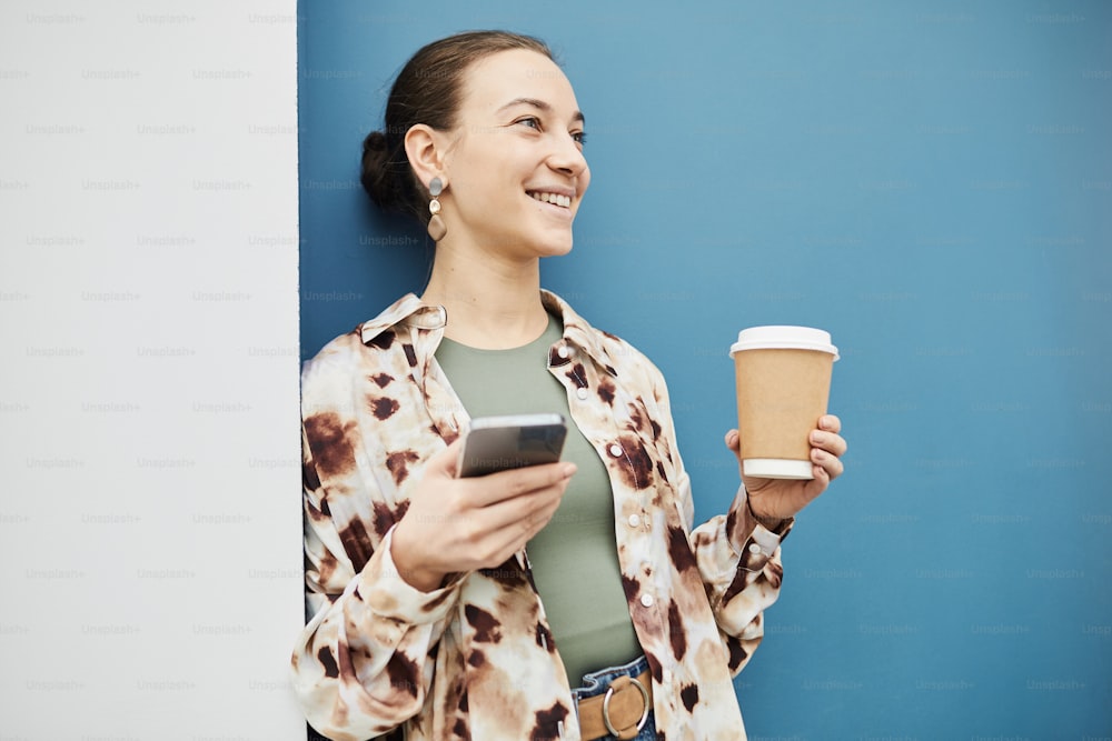 Vibrant minimal portrait of smiling young woman holding coffee cup against blue wall with candid smile, copy space