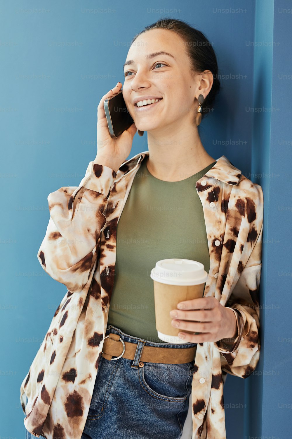 Vibrant portrait of young woman talking by smartphone while leaning casually against blue wall with genuine smile