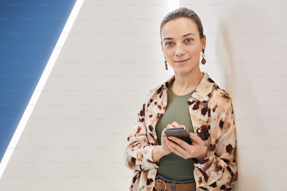 Graphic minimal portrait of smiling young woman holding phone and looking at camera by white wall in office, copy space