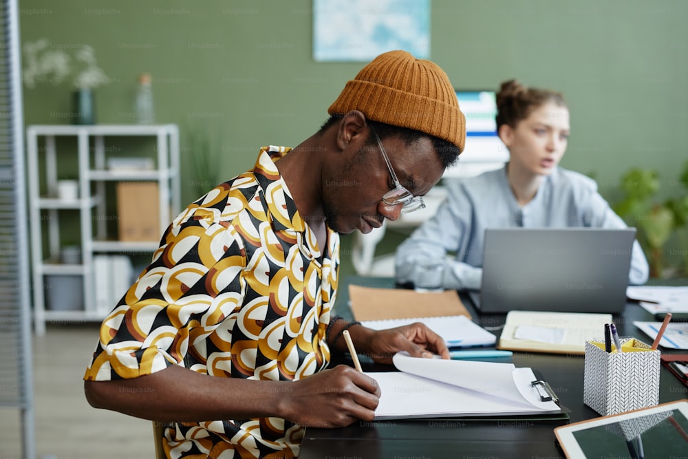 Vista lateral de un joven negro tomando notas en una reunión de negocios con el equipo creativo en la oficina