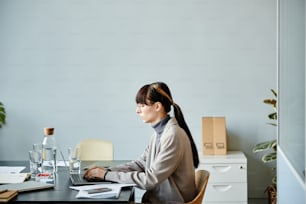Vista lateral mínima Retrato de una mujer joven usando la computadora en la mesa de reuniones en la oficina contra la pared azul pálido, espacio de copia