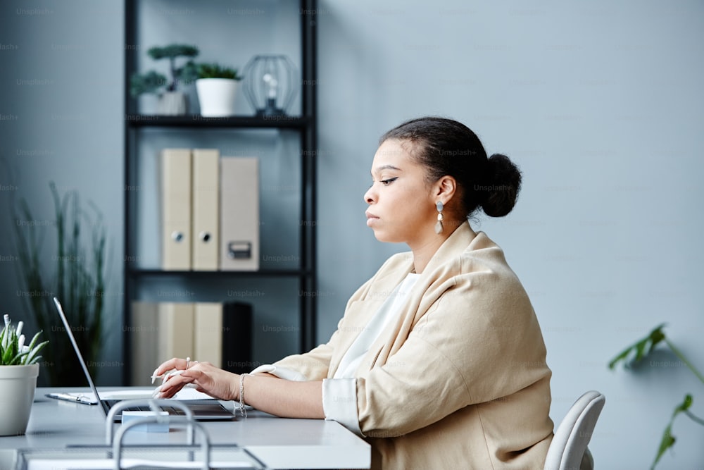 Minimal side view portrait of young black businesswoman using laptop at worplace in office against grey background