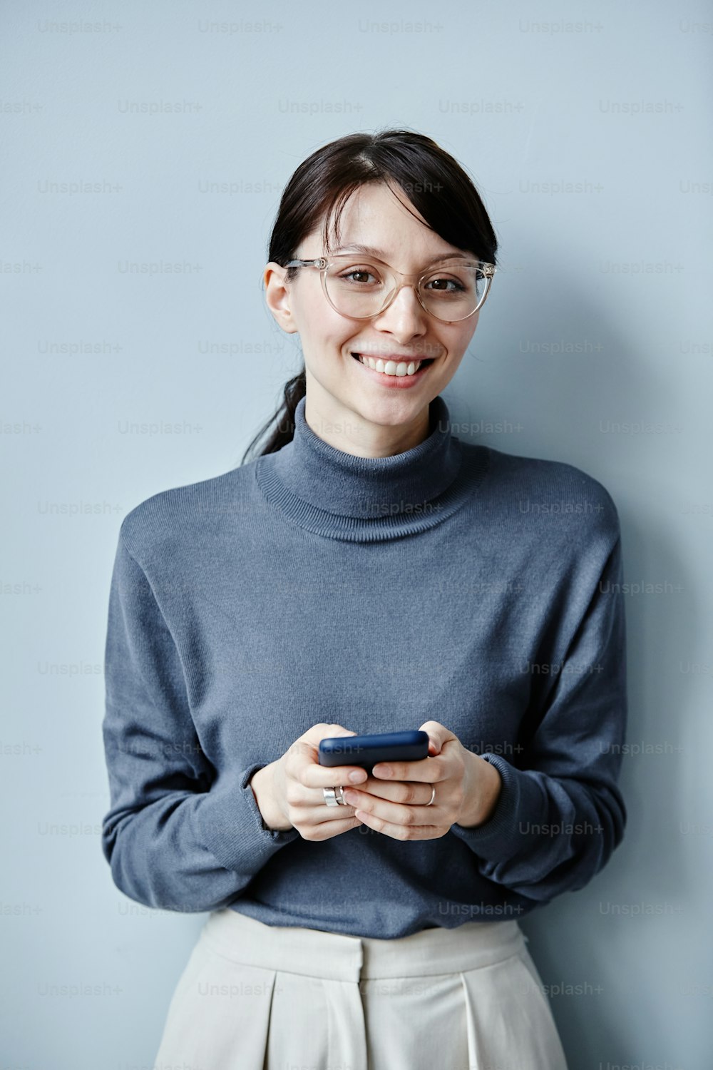 Candid portrait of young businesswoman holding smartphone and smiling at camera against simple blue background
