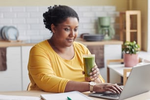 Porträt einer übergewichtigen schwarzen Frau, die Smoothie trinkt und zu Hause einen Laptop benutzt