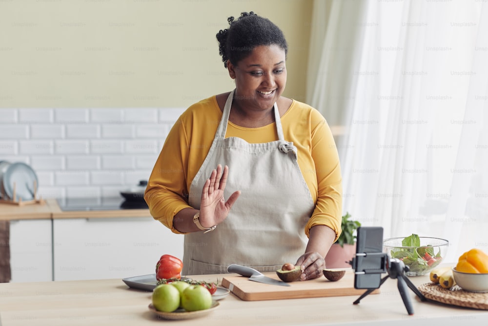 Portrait of young black woman cooking healthy meal in kitchen and waving at camera during livestream, copy space
