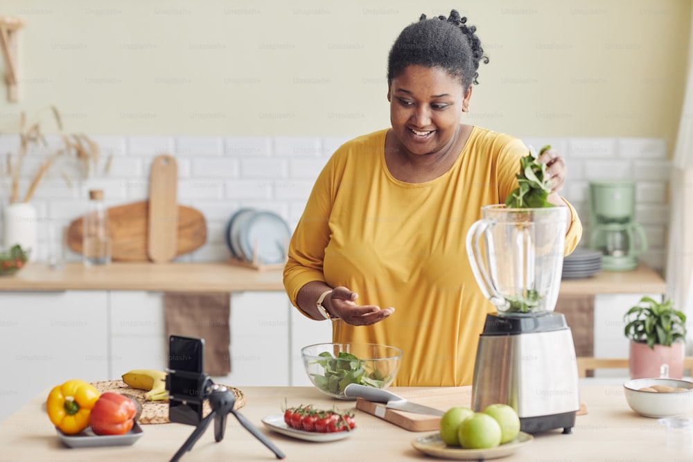 Ritratto di donna nera sorridente che usa il frullatore mentre prepara un pasto sano in cucina e filma video di cucina, spazio di copia