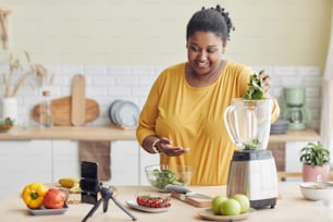 Portrait d’une femme noire souriante utilisant un mélangeur tout en préparant un repas sain dans la cuisine et en filmant une vidéo de cuisine, espace de copie