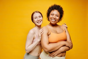 Portrait à la taille de deux jeunes femmes souriantes à la peau foncée et à la peau claire s’embrassant tout en se tenant debout sur un fond jaune vif en sous-vêtements